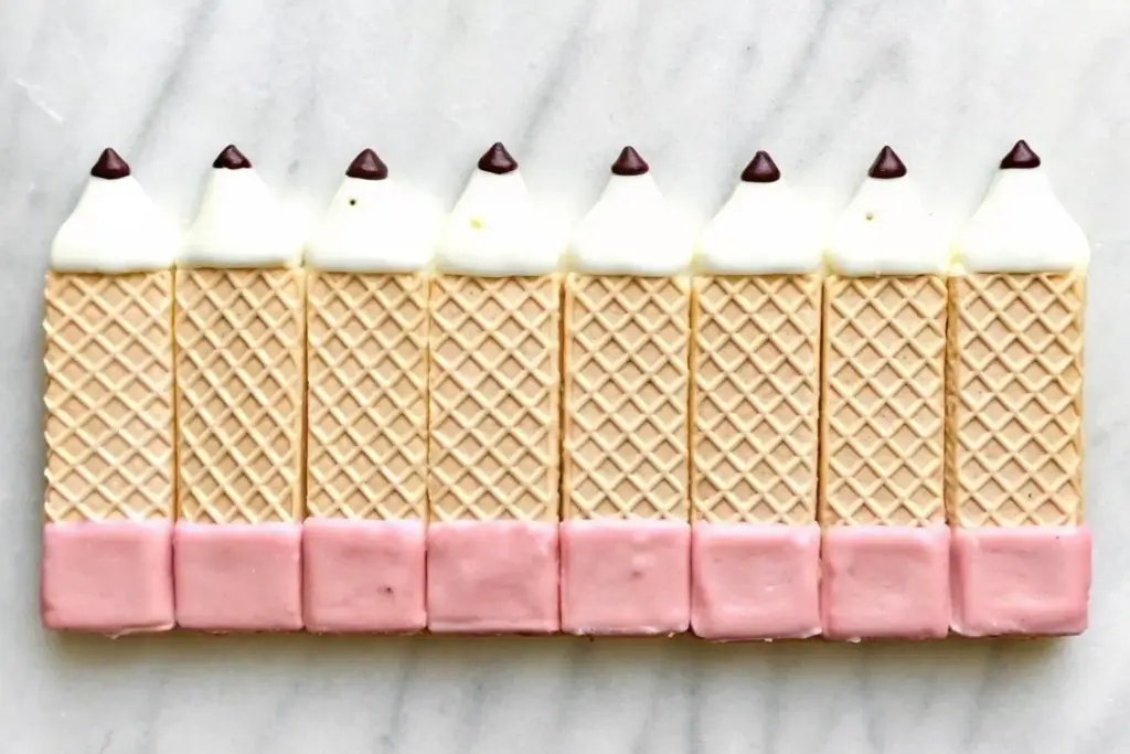 Five wafer pencil cookies with pink chocolate erasers, white chocolate tips, and mini chocolate chip leads arranged in a straight row on a marble surface.