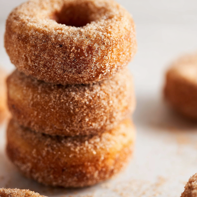 A stack of pumpkin spice donuts coated in cinnamon sugar, with a soft, golden texture and one donut placed on a light background.