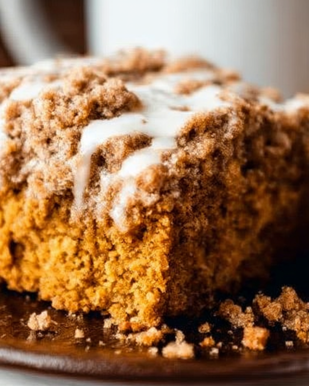 Pumpkin_Coffee_Cake_Recipe_natural A thick slice of pumpkin coffee cake topped with a crumbly cinnamon streusel and drizzled with white icing, sitting on a dark plate with crumbs scattered around.