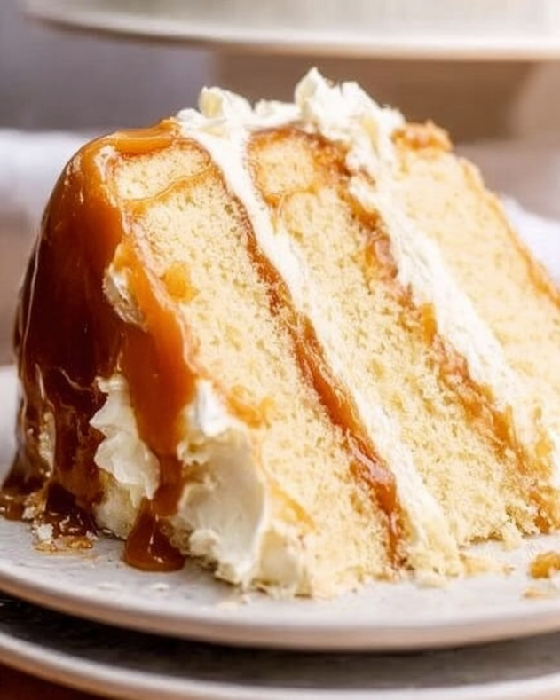 Slice of fluffy vanilla layer cake with creamy white frosting, generously drizzled with golden salted caramel sauce, highlighting smooth layers and a rich, gooey topping.​