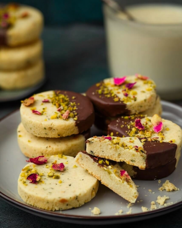 Almond and Pistachio Kulfi Shortbread cookies, some half-dipped in chocolate and garnished with chopped pistachios and dried rose petals, arranged on a plate with a glass of milk in the background