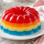 Rainbow layered gelatin dessert unmolded onto a white plate, showing red, orange, yellow, and blue stripes