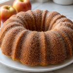Apple cider donut Bundt cake coated with cinnamon sugar on a white platter beside apples and cider.