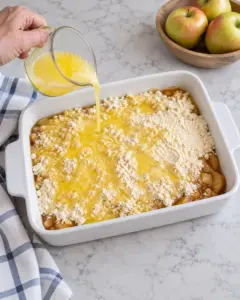 Melted butter being poured over dry cake mix layered above caramel apple pie filling.