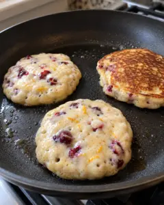 Cranberry orange ricotta pancakes cooking in a skillet with set bubbles at the edges before flipping